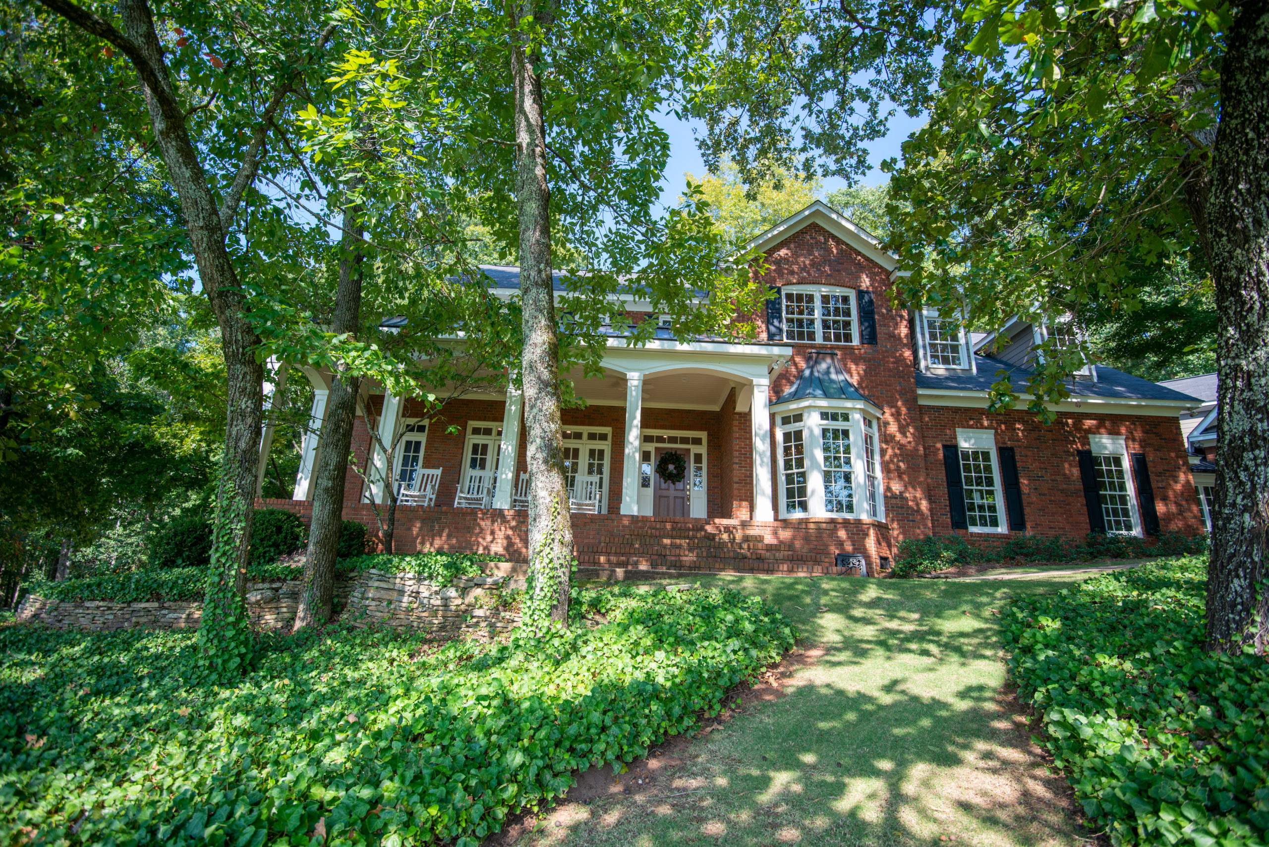 Red brick two-story house with white columns on the front porch, surrounded by mature trees and ivy ground cover on a sunny day.